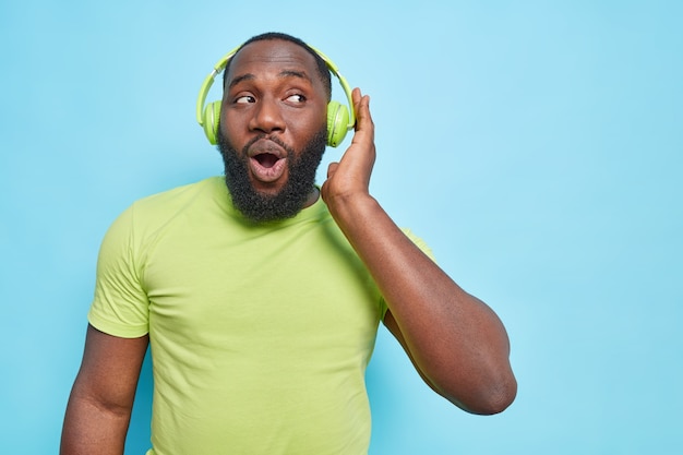 Un homme barbu curieux et surpris garde la main sur les écouteurs garde la bouche ouverte vêtu d'un t-shirt vert écoute la musique préférée isolée sur un mur bleu