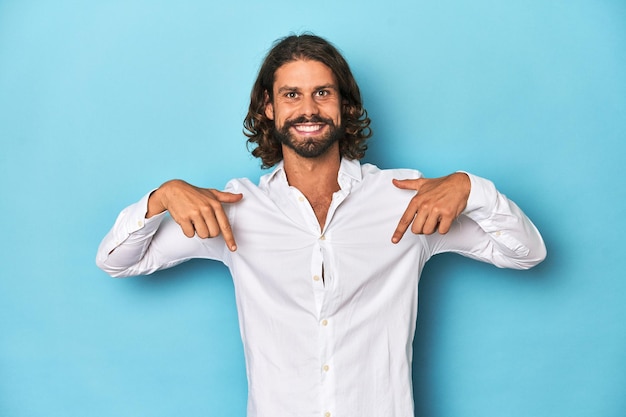 Homme barbu dans une chemise blanche fond bleu pointe vers le bas avec des doigts sentiment positif