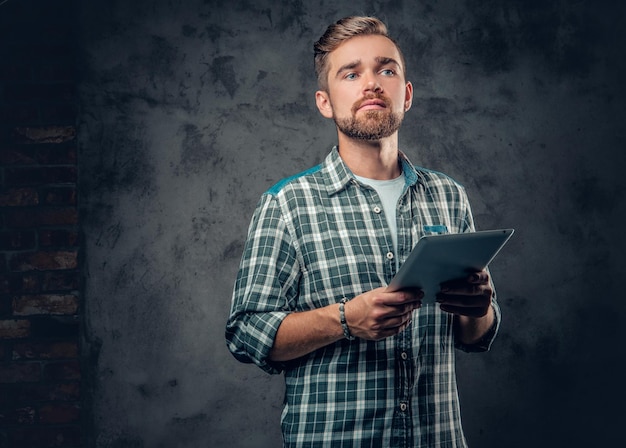 Un homme barbu dans une chemise polaire tient une tablette PC sur fond gris.