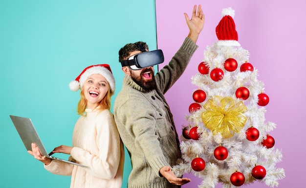 Homme barbu dans des lunettes virtuelles d et femme en bonnet de noel avec ordinateur portable près de l'arbre de noël