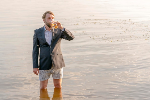Homme barbu dans une veste grise et des sous-vêtements avec une bière à la main debout jusqu'aux genoux dans l'eau