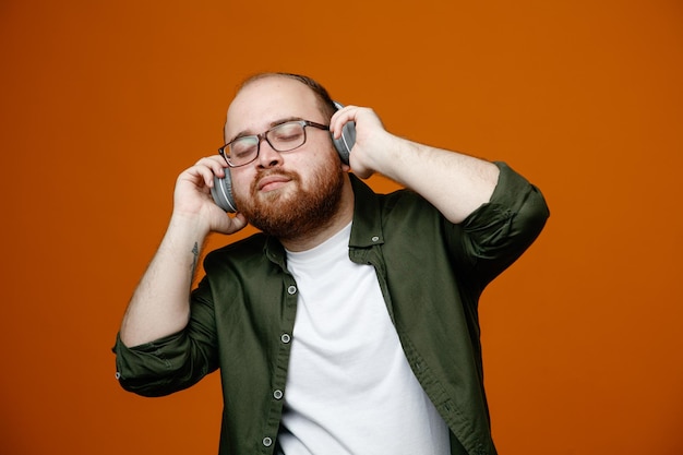 Homme barbu dans des vêtements décontractés portant des lunettes avec des écouteurs calme et détendu en écoutant de la musique debout sur fond orange