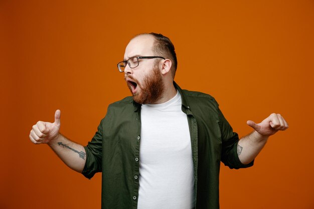 Homme barbu dans des vêtements décontractés portant des lunettes heureux et excité montrant les pouces vers le haut debout sur fond orange