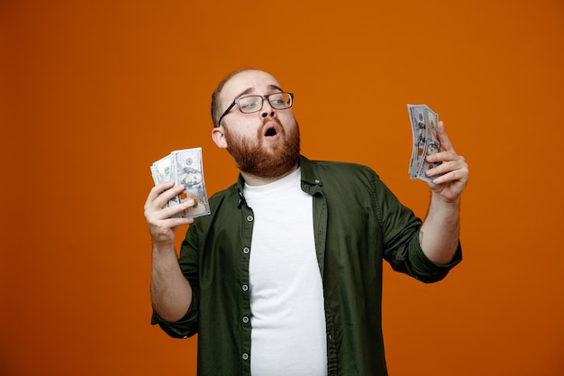 Homme barbu dans des vêtements décontractés portant des lunettes tenant de l'argent heureux et surpris debout sur fond orange