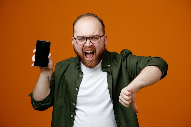 Homme barbu dans des vêtements décontractés portant des lunettes tenant un smartphone regardant la caméra en colère et frustré montrant le pouce vers le bas debout sur fond orange