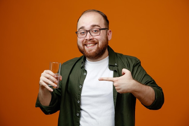 Homme barbu dans des vêtements décontractés portant des lunettes tenant un verre d'eau pointant avec l'index dessus souriant joyeusement heureux et positif debout sur fond orange