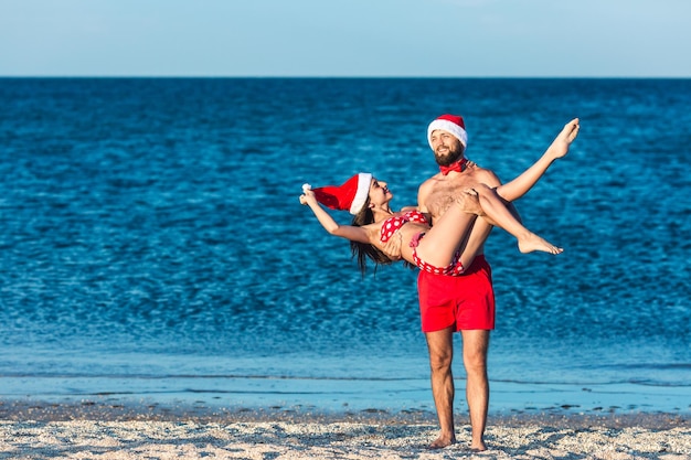 Homme barbu drôle et fille en chapeaux de Père Noël. le gars tient la fille dans ses bras. Noël d'été sur la plage de la mer.