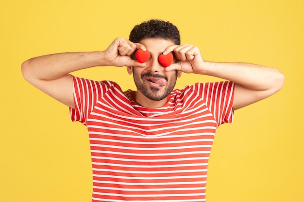 Photo homme barbu drôle en t-shirt rayé s'amusant tenant le coeur de jouet près des yeux et montrant la langue, confession d'amour, célébration de la saint valentin. tourné en studio intérieur isolé sur fond jaune