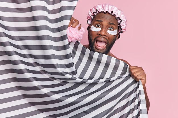 Un homme barbu effrayé émotionnel se cache derrière un rideau de douche ne s'attendait pas à ce que quelqu'un entre dans la salle de bain porte un chapeau imperméable et des patchs de collagène isolés sur un mur rose