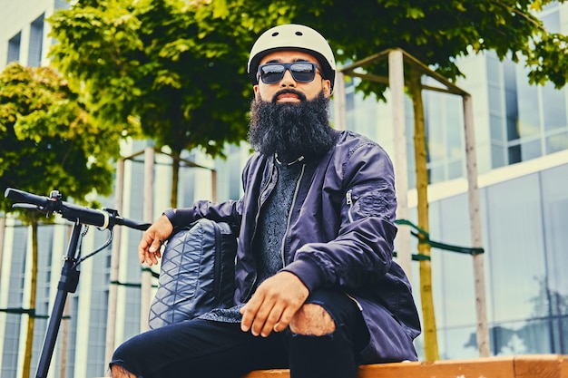 Homme barbu élégant en lunettes de soleil se reposant sur un banc après avoir roulé en scooter électrique au centre-ville.