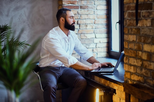 Un homme barbu élégant travaille avec un ordinateur portable dans une pièce avec un intérieur loft.