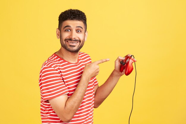 Homme barbu enfantin positif en t-shirt rayé pointant le doigt sur le joystick filaire regardant la caméra avec une expression heureuse accro aux jeux virtuels Studio intérieur tourné isolé sur fond jaune