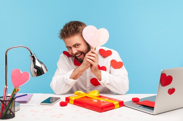 Homme barbu espiègle assis sur le lieu de travail tout couvert de bâtons de coeur et souriant, tenant une carte de voeux rose en forme de coeur, préparant un cadeau. Tourné en studio intérieur isolé sur fond bleu