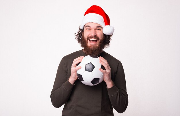 Homme barbu étonné tenant un ballon de football et criant et portant un chapeau de père Noël