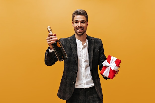 Un homme barbu excité dans un pantalon à carreaux et une chemise blanche sourit se penche sur la caméra et tient une bouteille de champagne et une boîte-cadeau rouge sur fond orange