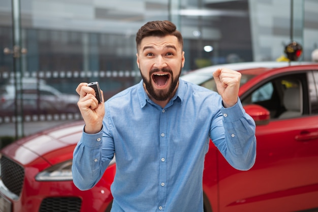 Homme barbu excité hurlant joyeusement, tenant les clés de sa nouvelle voiture.