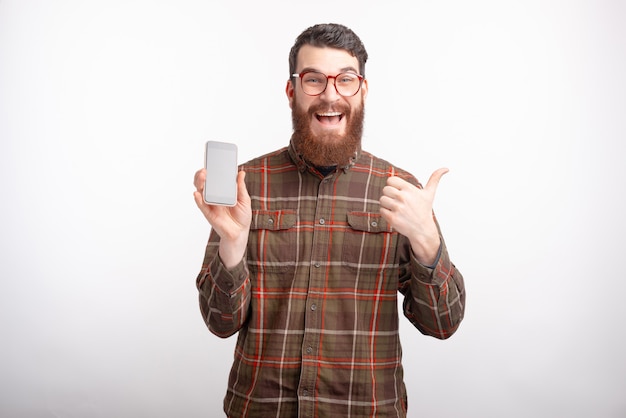 Un homme barbu excité montre comme un geste et son téléphone sur fond blanc.