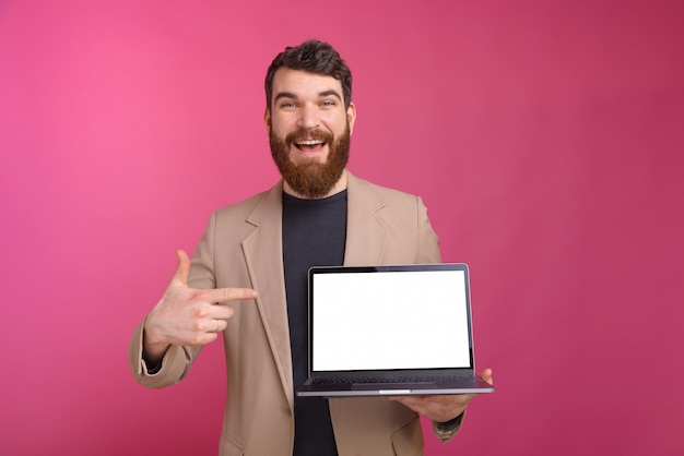 Un homme barbu excité pointe son ordinateur portable qu'il tient sur fond rose.