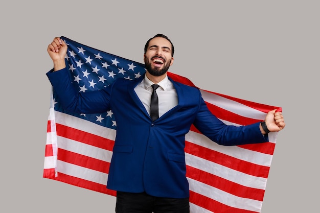 Homme barbu excité tenant le drapeau américain et regardant la caméra avec un regard de joie célébrant la fête nationale portant un costume de style officiel Prise de vue en studio intérieure isolée sur fond gris