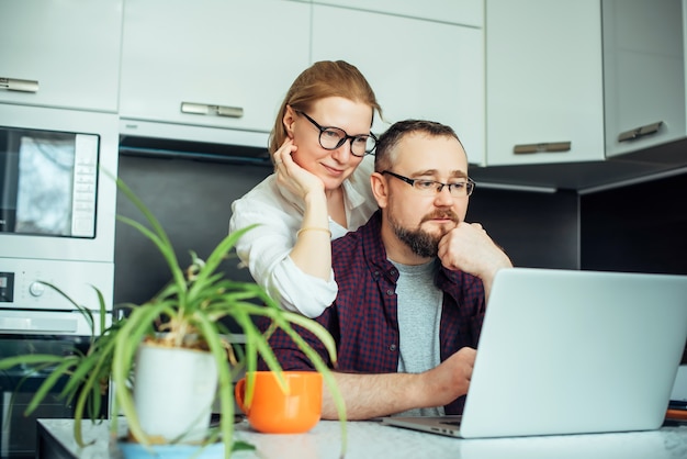 Homme barbu et femme avec des lunettes dans la cuisine regardant un ordinateur portable, discutant des nouvelles du matin. Couple d'adultes passant du temps ensemble à la maison.