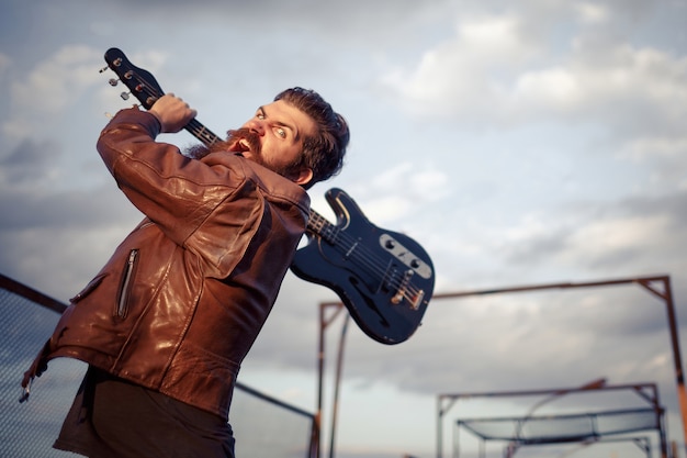 Homme Barbu Fou Aux Cheveux Gris Dans Une Veste En Cuir Marron Brandit Une Guitare électrique Noire Contre Le Ciel