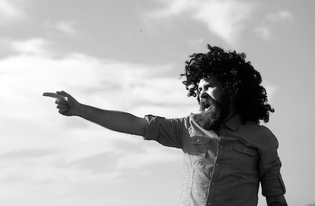 Un homme barbu fou et drôle avec une perruque noire montrant le doigt sur le fond du ciel des expressions faciales drôles