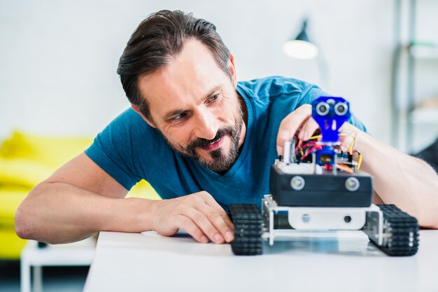 Homme barbu fou de joie assis à la table tout en construisant un robot à la maison