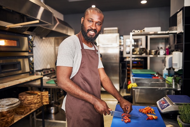 homme barbu gai en tablier travaillant avec un couteau pendant la cuisson des plats