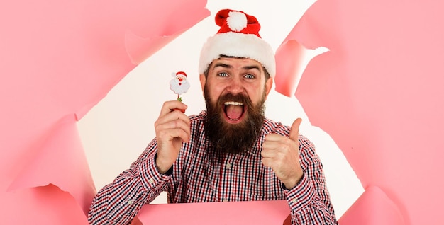 Un homme barbu heureux dans un chapeau de Père Noël avec une petite sucette de bonbons de Père Noëls montrant le pouce en l'air en regardant à travers