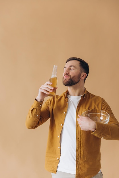 Un homme barbu heureux et souriant en chemise jaune buvant de la bière et mangeant des frites sur un fond beige