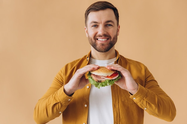 Un homme barbu heureux et souriant en chemise jaune mangeant un hamburger sur un fond beige