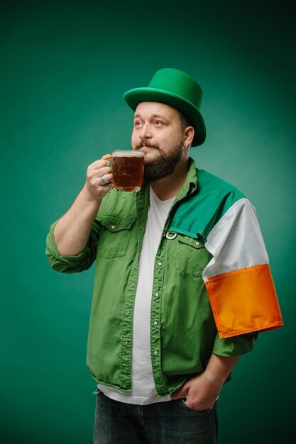 Photo homme barbu heureux avec un verre de bière et un drapeau irlandais sur un fond vert foncé jour de saint-patrick