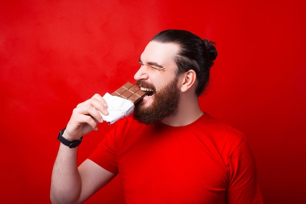 Homme barbu hipster mangeant une savoureuse barre de chocolat sur mur rouge