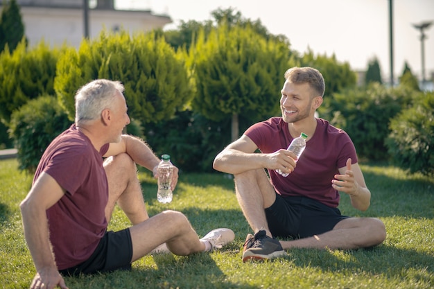 Homme barbu et homme aux cheveux gris assis sur l'herbe, discuter, boire de l'eau