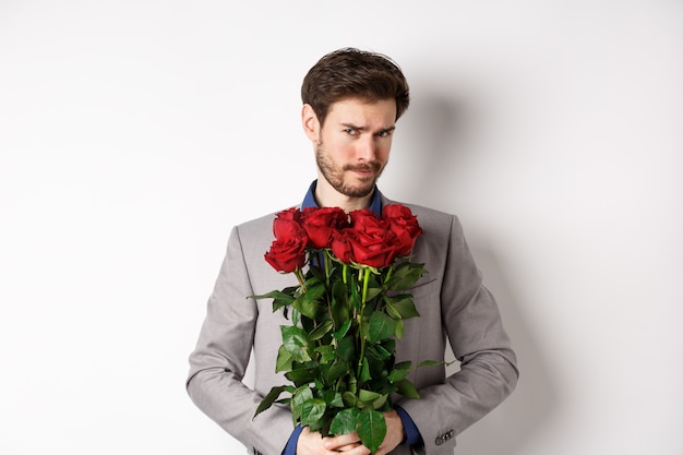 Homme barbu indécis en costume, tenant des fleurs roses et regardant la caméra incertain, allant à un rendez-vous romantique, debout sur fond blanc.