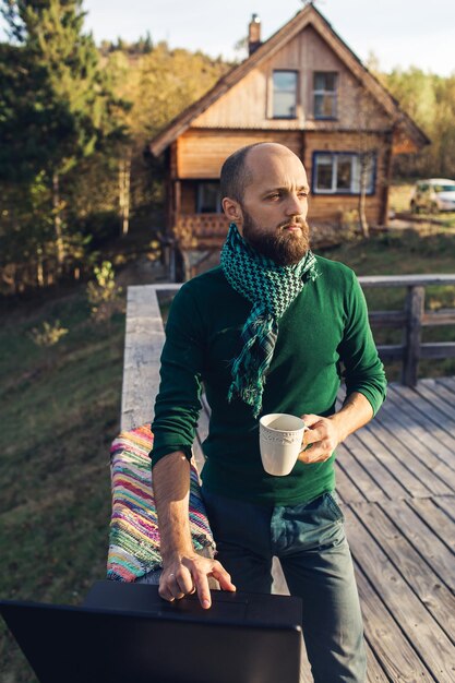 Un homme barbu indépendant est debout sur une plate-forme dans les montagnes avec un ordinateur portable et boit du café.