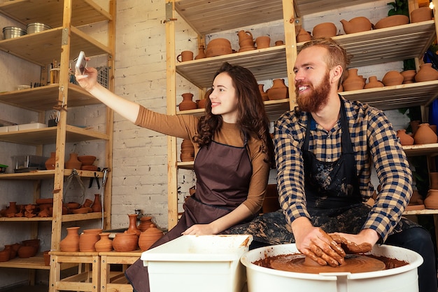 Un homme barbu et une jeune femme moulent un vase d'argile sur un tour de potier et réalisent un selfie au téléphone dans un atelier de poterie. potier, argile, vase, atelier de poterie. maître et élève.