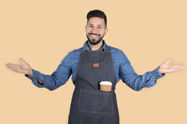 Homme barbu latino hispanique avec tablier bleu se tient les bras grands ouverts invitant à passer très souriant sur fond jaune