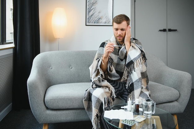 Homme barbu malade qui a un mauvais rhume ou une grippe saisonnière assis sur un canapé à la maison Un gars avec de la fièvre portant un plaid chaud frissonnant avec une expression de visage inquiet