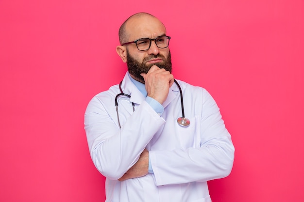 Homme barbu médecin en blouse blanche avec stéthoscope autour du cou portant des lunettes regardant de côté avec la main sur son menton pensant avec un visage sérieux debout sur fond rose