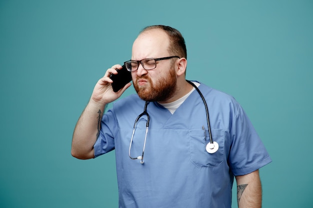 Homme barbu médecin en uniforme avec stéthoscope autour du cou portant des lunettes à mécontent tout en parlant au téléphone mobile debout sur fond bleu