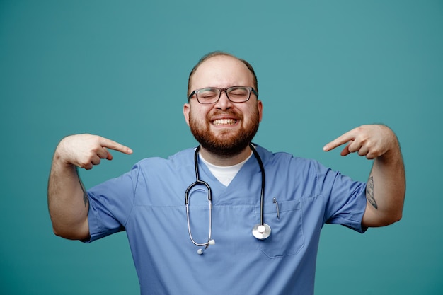 Homme barbu médecin en uniforme avec stéthoscope autour du cou portant des lunettes satisfait souriant largement pointant vers lui-même debout sur fond bleu