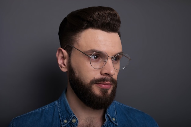 Homme barbu moderne et intelligent confiant dans des vêtements en denim décontractés et des lunettes pose