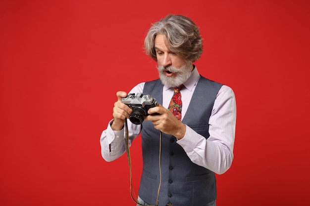 Homme barbu à moustache aux cheveux gris âgé étonné en chemise classique gilet cravate colorée isolé sur fond rouge en studio. Concept de style de vie des gens. Maquette de l'espace de copie. Tenez un appareil photo vintage rétro.