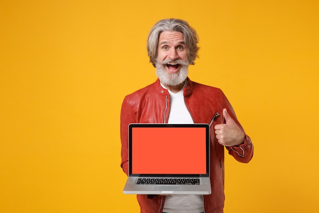 Homme barbu à moustache aux cheveux gris âgé et excité en veste de cuir isolé sur fond jaune. Concept de style de vie des gens. Maquette de l'espace de copie. Tenez un ordinateur portable avec un écran vide montrant le pouce vers le haut.