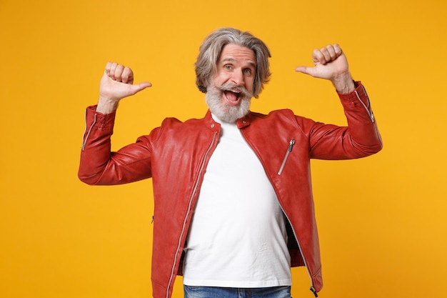 Photo homme barbu à moustache aux cheveux gris âgé et excité en veste de cuir rouge posant isolé sur fond orange jaune en studio. concept de style de vie des gens. maquette de l'espace de copie. pointant les pouces sur lui-même.