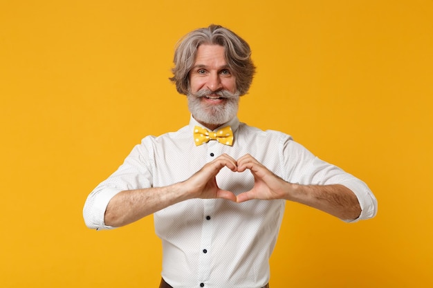 Homme barbu à moustache aux cheveux gris souriant âgé en chemise blanche et noeud papillon posant isolé sur fond orange jaune. Concept de style de vie des gens. Maquette de l'espace de copie. Montrant le coeur de forme avec les mains.