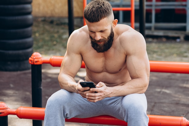 Homme barbu musclé fort à la recherche de vidéo sur smartphone après une formation dans un gymnase de rue en plein air. Concept de mode de vie d'entraînement.