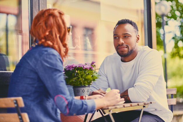 Un homme barbu noir et une femme rousse boivent du café dans un café dans une rue.