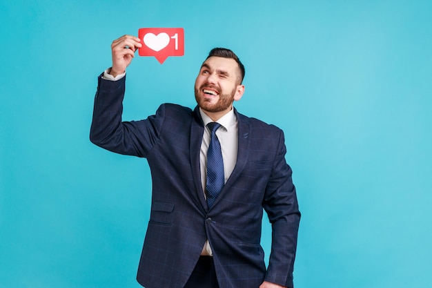 Homme barbu optimiste vêtu d'un costume de style officiel tenant le réseau social icône Heart Like sur la tête en regardant le symbole et en faisant un clin d'œil Prise de vue en studio intérieure isolée sur fond bleu